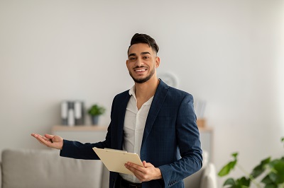 Friendly arab male psychologist with clipboard showing something with his hand at mental health clinic, free space. Cheerful psychotherapist posing and smiling at office